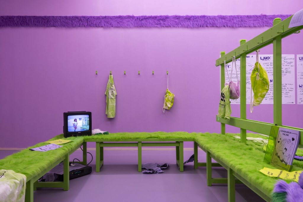 In this shot, we see only a portion of the fur-covered bench. On the bench is an old 90s era thick black television set, which features the still of a white girl dancing on it. Behind the TV and bench are green hooks on the lilac wall, holding a backpack and green tie-dyed T-shirt.