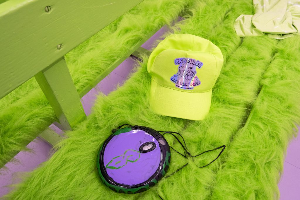 This is a close-up of the discman, which has a green middle-school style ‘s’ painted into a purple background on its face. Nearby is a green hat with the ‘axe pulse wrestling club’ logo.