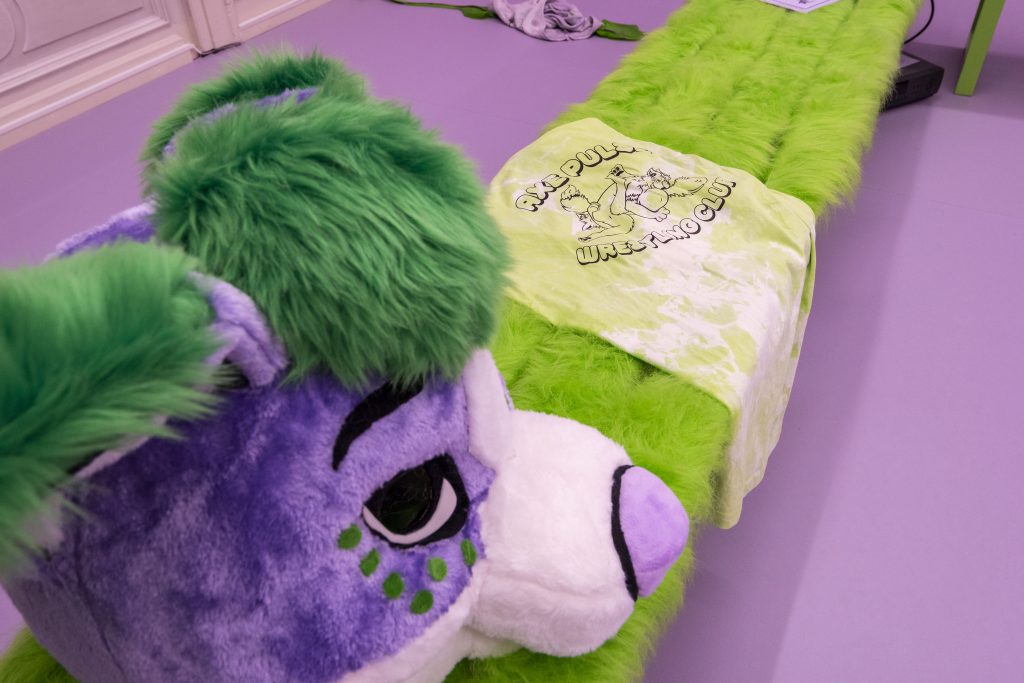This is a close-up of the mascot team furry head near a t shirt that has been draped over the bench. The head looks like some kind of purple and green fox or dog, with a white nose and cute pink nose.