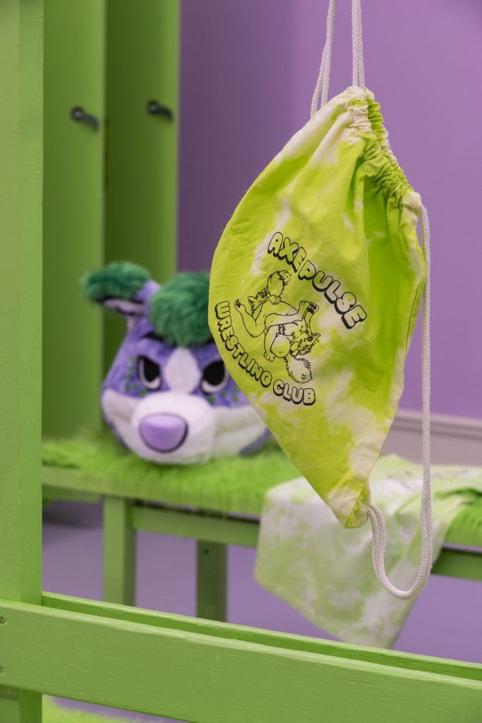 This is a close-up of a green tie-dyed backpack with the ‘AXE PULSE wrestling club’, which features two foxes wrestling each other, each with their noses erotically close to each other’s buttholes. Behind it we see the mascot head sitting on the bench.