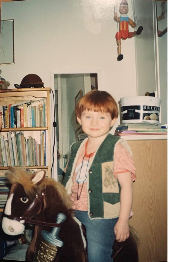Image of Flip around the age of four, riding on Bobby the toy horse and wearing his favorite orange striped shirt. Slung around Bobby's neck there is a plastic dagger in sheath.