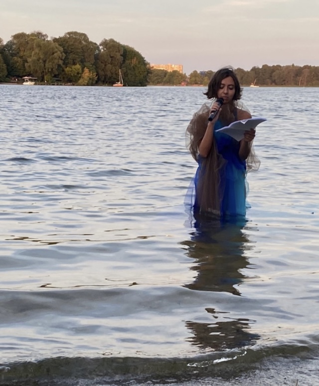Daniela can be seen holding a microphone and paper. She is knee deep in water, reading. Her outfit is dark blue, light blue, and brown gauzy material floating on the water.