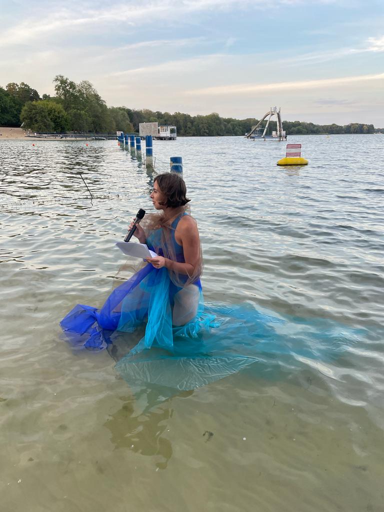 A side view of Daniela.  Her gauzy outfit spreads out through the water.  In the background a beach for swimming can be seen.