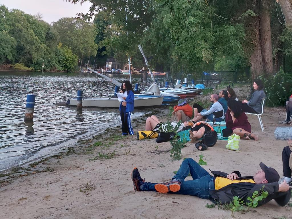 Lorena Juan Gutierrez, a white-skinned woman with long black curly hair, is holding a microphone and paper and reading to an audience of 10 people lying and sitting on the ground.  Lorena is wearing a long blue shirt, short white tank top, and blue track suit pants.  She is standing at the shoreline of a lake, with boats, trees, and water behind her. 