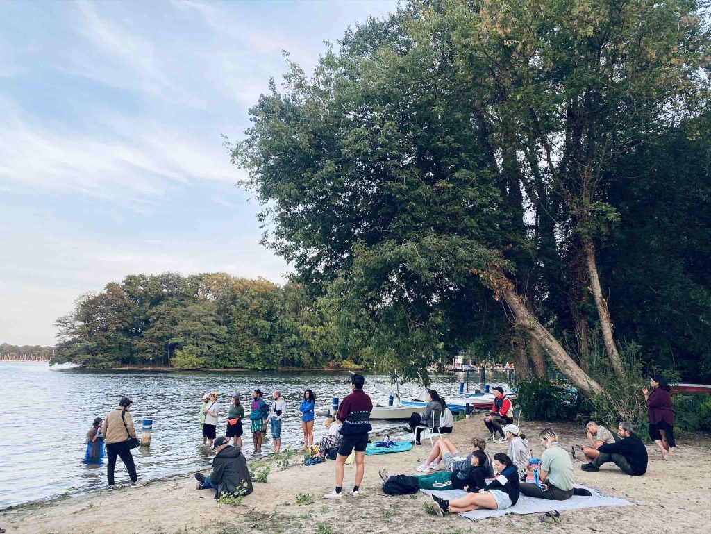 wide view of a large leannig over the audience and daniela at the shore.  The beach, water, and forest stretch out around them.