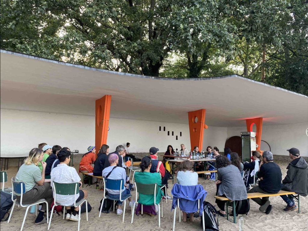 A group of 20 people sit watching artists on a panel outside.  The artists are elft to right Meghna Singh, Zinzi Buchanan, Frances Breden, Daniela Medina Poch, Harley Aussoleil, and Lorena Juan Gutierrez.