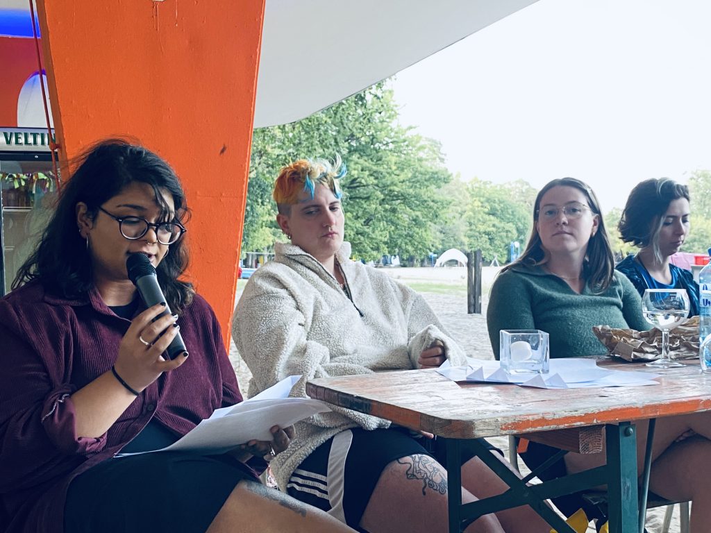 A close up of Meghna, a brown-skinned woman with long dark hair and round black glasses and a dark purple shirt.  She is holding the microphone and reading.  Zinzi, a white-skinned person with short orange and blue hair and a white fleece jacket, is listening.  Frances, a white-skinned woman with ling brown hair, round glasses, and a green long shirt is looking into the camera.  Daniela, a white-skinned woman with a brown bob and blob shirt, is looking down.  Water bottles, wine glasses, and papers are on the table in front of them all.