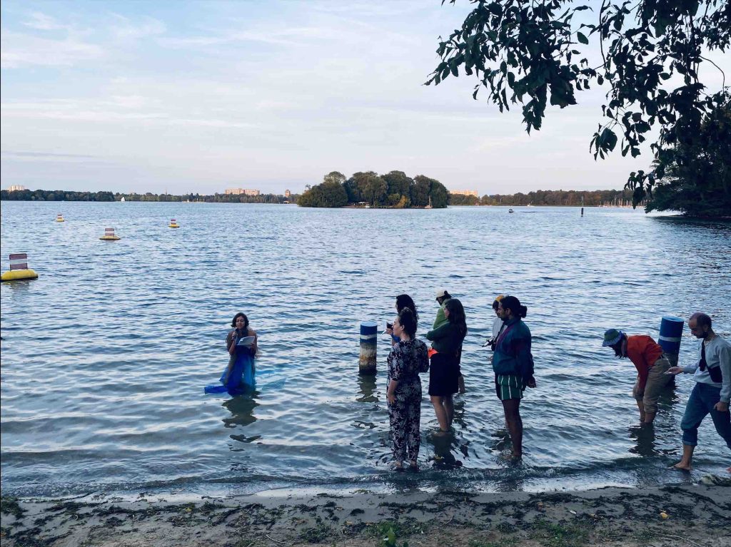 10 audience members stand with their feet in the water listening to Daniela.  The shining water and the shoreline in the distance can be seen.