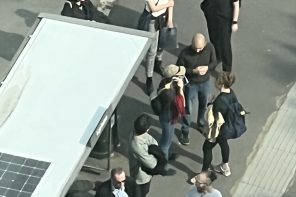 This looks like a photo that’s been edited to appear almost drawn. Featured is a group of people waiting at some kind of bus stop-shelter. The photo is shot from above, like from a drone or the window of an apartment overlooking the stop. Of the people there, you can clearly make out a man talking to two women.