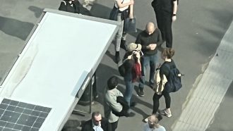 This looks like a photo that’s been edited to appear almost drawn. Featured is a group of people waiting at some kind of bus stop-shelter. The photo is shot from above, like from a drone or the window of an apartment overlooking the stop. Of the people there, you can clearly make out a man talking to two women.