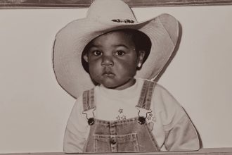 This image features D'orjay as a baby. They wear a cowboy hats and dungarees.