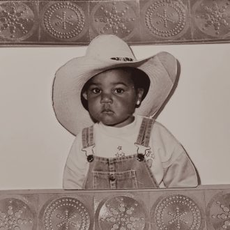 This image features D'orjay as a baby. They wear a cowboy hats and dungarees.