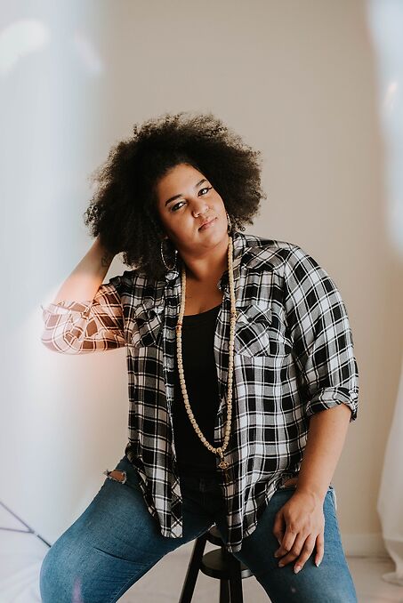 Here is an image of D’orjay The Singing Shaman smiling and sitting on a black stool. They wear ripped blue jeans, a black t-shirt and a black and white flannel, hoop earrings and a long beaded necklace. One hand rests on their thigh, the other by their head.